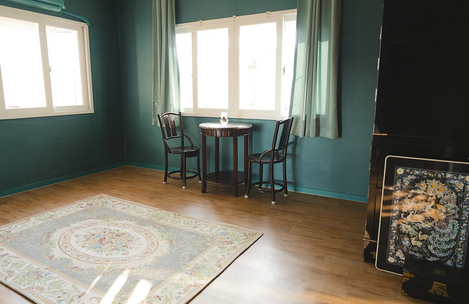 A teal room at Botonglee Studio with windows on two walls, a round table and two chairs
